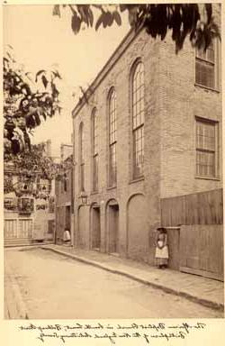 <p>Sepia photograph of a street and the side of a tall, brick building with four arched windows and doors and a large iron lamp. To the left of the building, a Black woman with a shawl, hat, apron, and long-sleeved dress stands against a wall with her hands clasped together in front of her.</p>