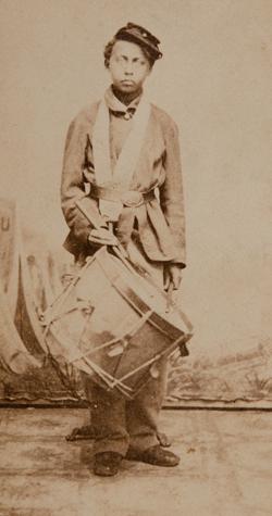 <p>A black and white photograph of a young, Black boy wearing a cap and a large army coat cinched at the waist with a belt standing against a white background. He holds one drumstick in each hand, one of which is resting against a large snare drum which hangs from his neck.</p>